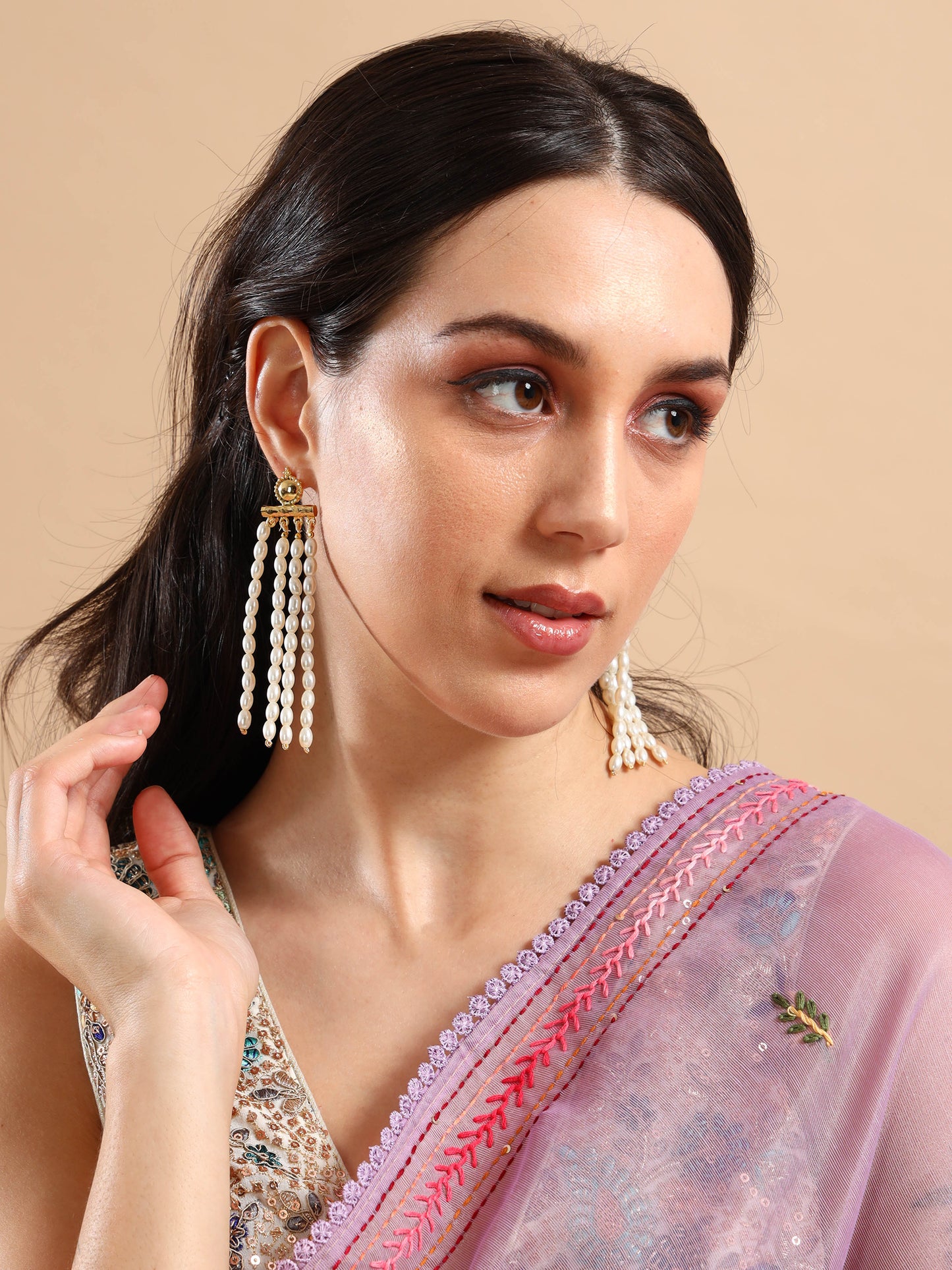 A woman in a light purple saree with pink embroidery looks to the side. She wears Pearl Rain Earrings—long, 24K gold plated earrings with pearls—and has dark, wavy hair pulled back and subtle makeup with soft eyeshadow and nude lipstick.