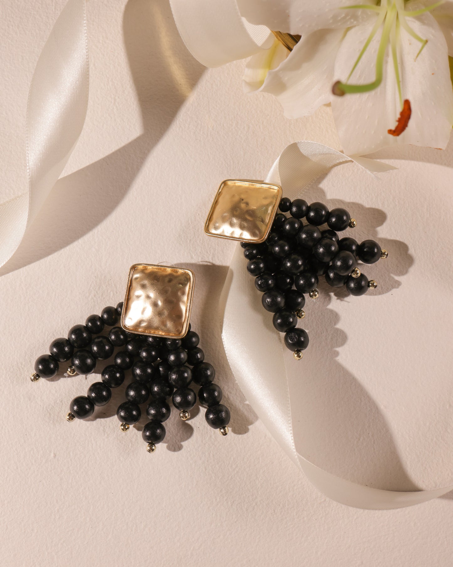Beady Black and Gold Earrings: gold-plated square stud earrings with dangling black bead clusters, shown on a light backdrop with white ribbon and lily—unique standout summer jewelry.