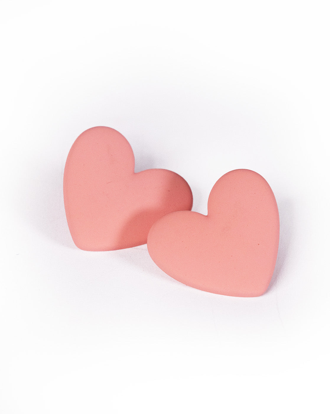 Two Love Hearts Earrings, pink and heart-shaped, rest on a white surface leaning together. Their smooth pastel finish gleams softly, evoking the delicate shine of elegant jewelry.