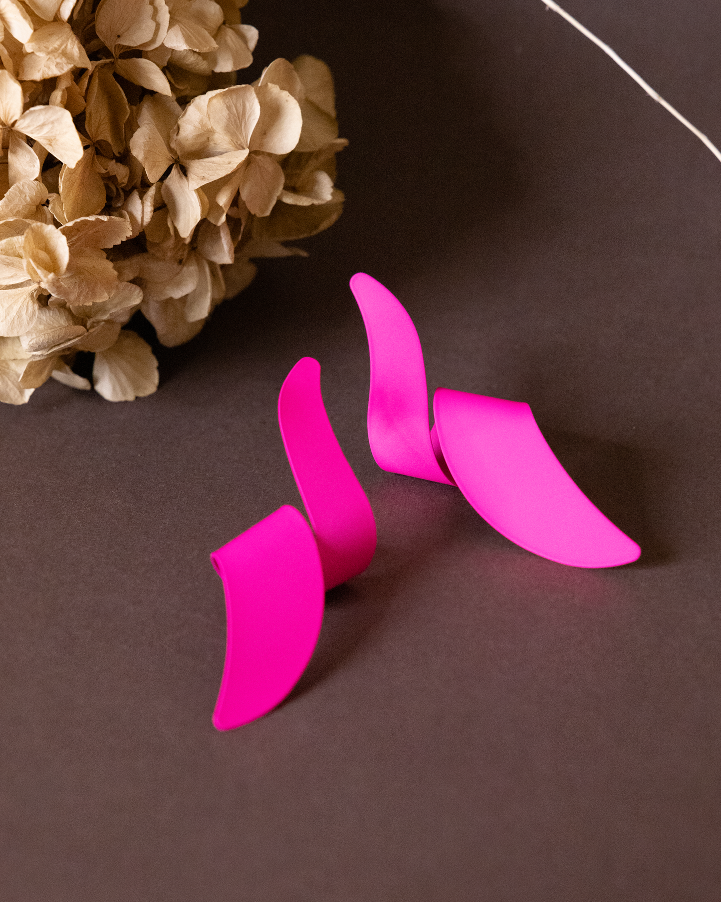 Four bright pink long twisted dangler earrings are arranged on a dark surface next to a cluster of dried beige hydrangea flowers.
