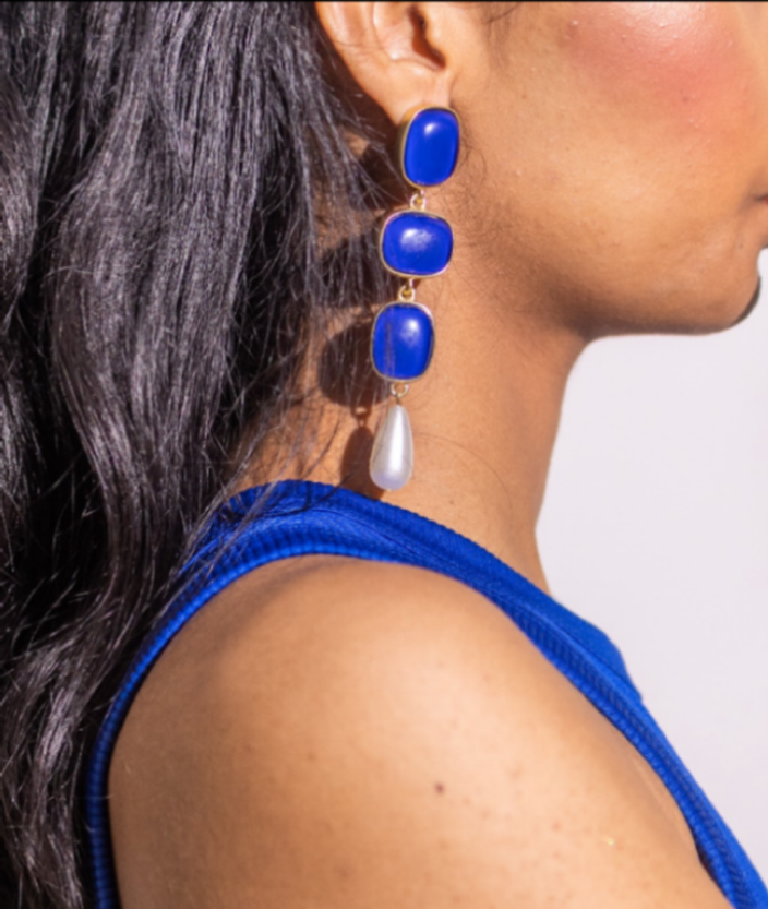 A person with long dark hair wears a bright blue sleeveless top and Blue Enamel Danglers—statement earrings with three blue stones and a dangling white teardrop-shaped bead.