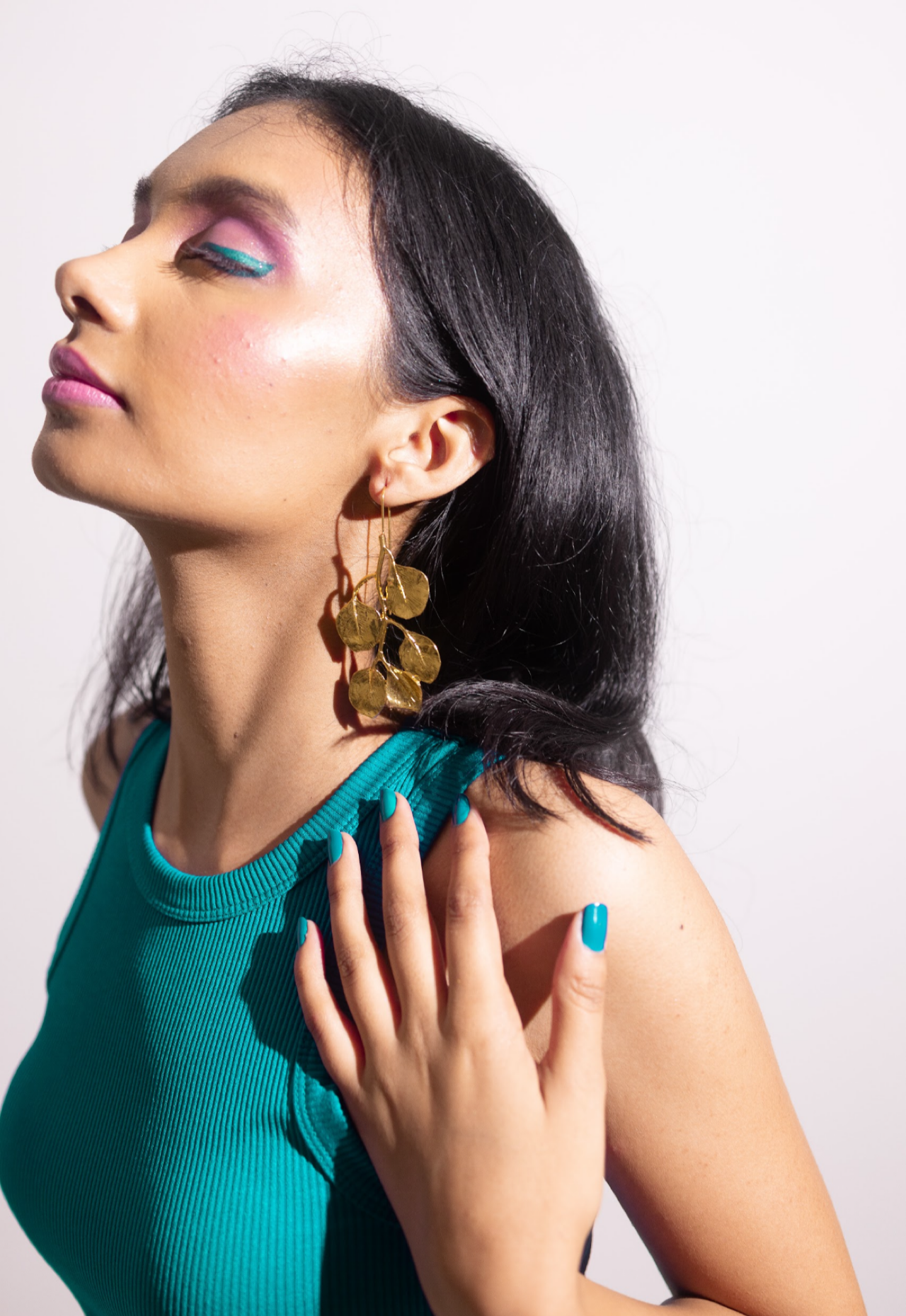 A woman with long dark hair wears Hanging Leaves Danglers, teal nail polish, and a teal sleeveless top. Eyes closed to reveal colorful makeup, she poses with one hand on her shoulder against a plain background.