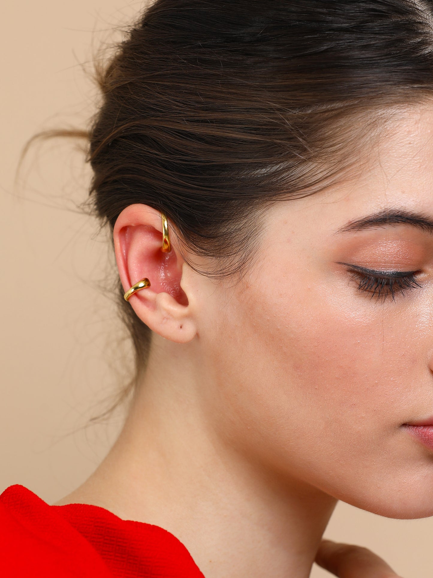 Close-up of a woman's ear adorned with a Gold twin ear cuff and gold hoop earrings, her brown hair tied back, wearing a red top against a beige background.