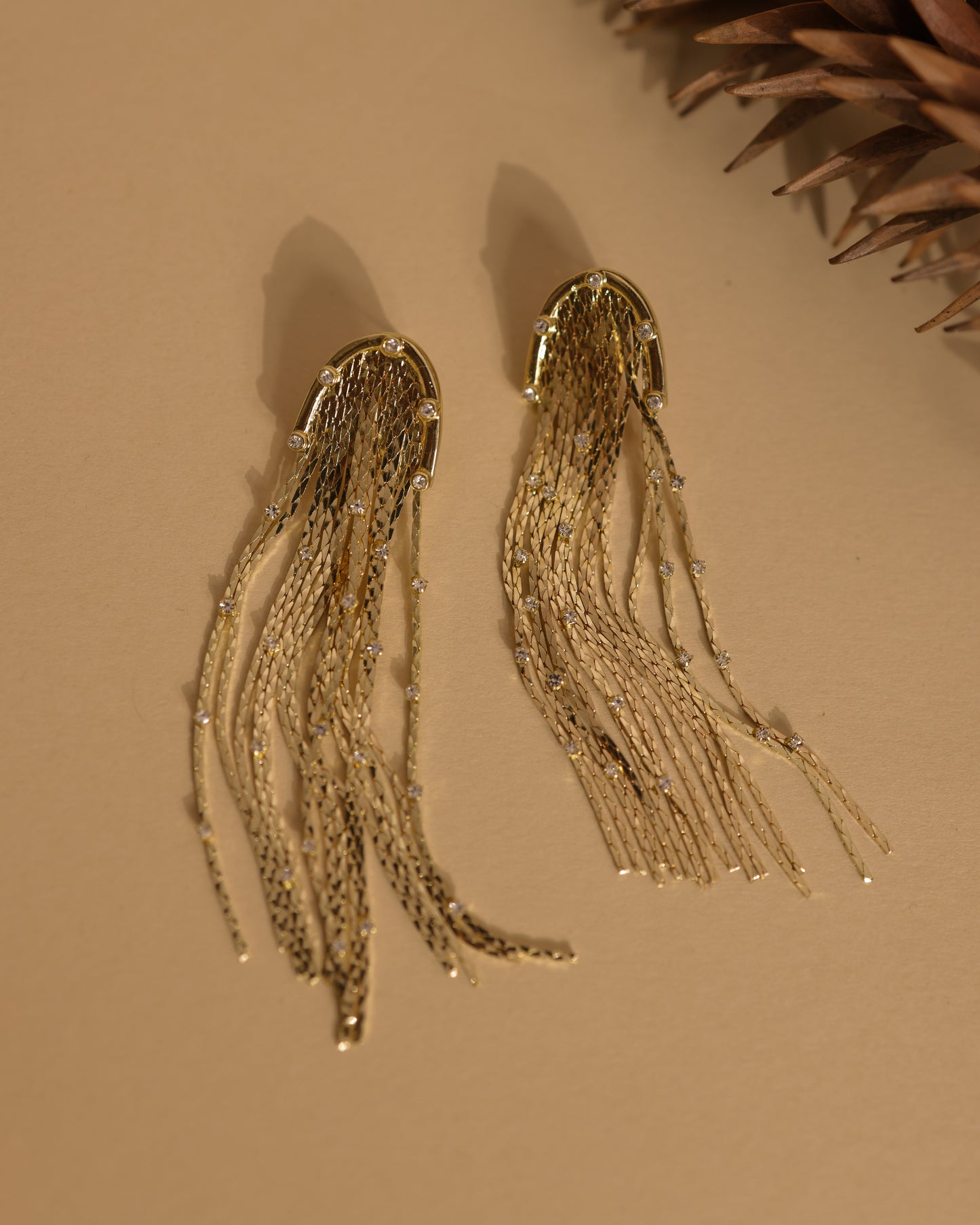 A pair of "Tassels With Small Zirconia" gold-tone earrings featuring cascading chain strands are displayed on a neutral beige surface, with a pinecone partially visible in the top right corner.