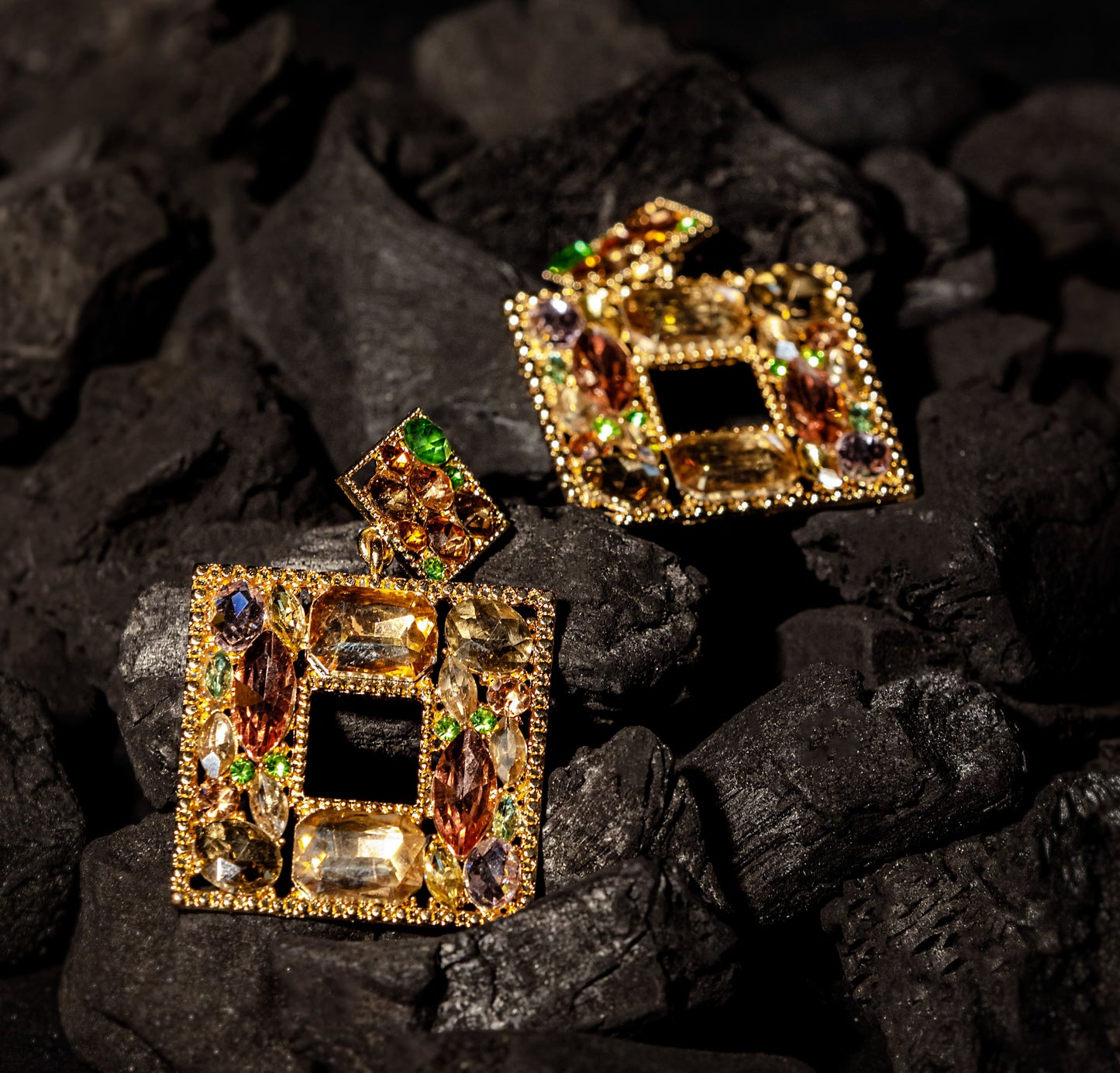 The Bella Earrings are ornate, square-shaped dangle earrings plated in 24K gold and adorned with green, brown, yellow, and clear gemstones for a vibrant, luxurious look. Displayed elegantly on a bed of dark charcoal pieces.