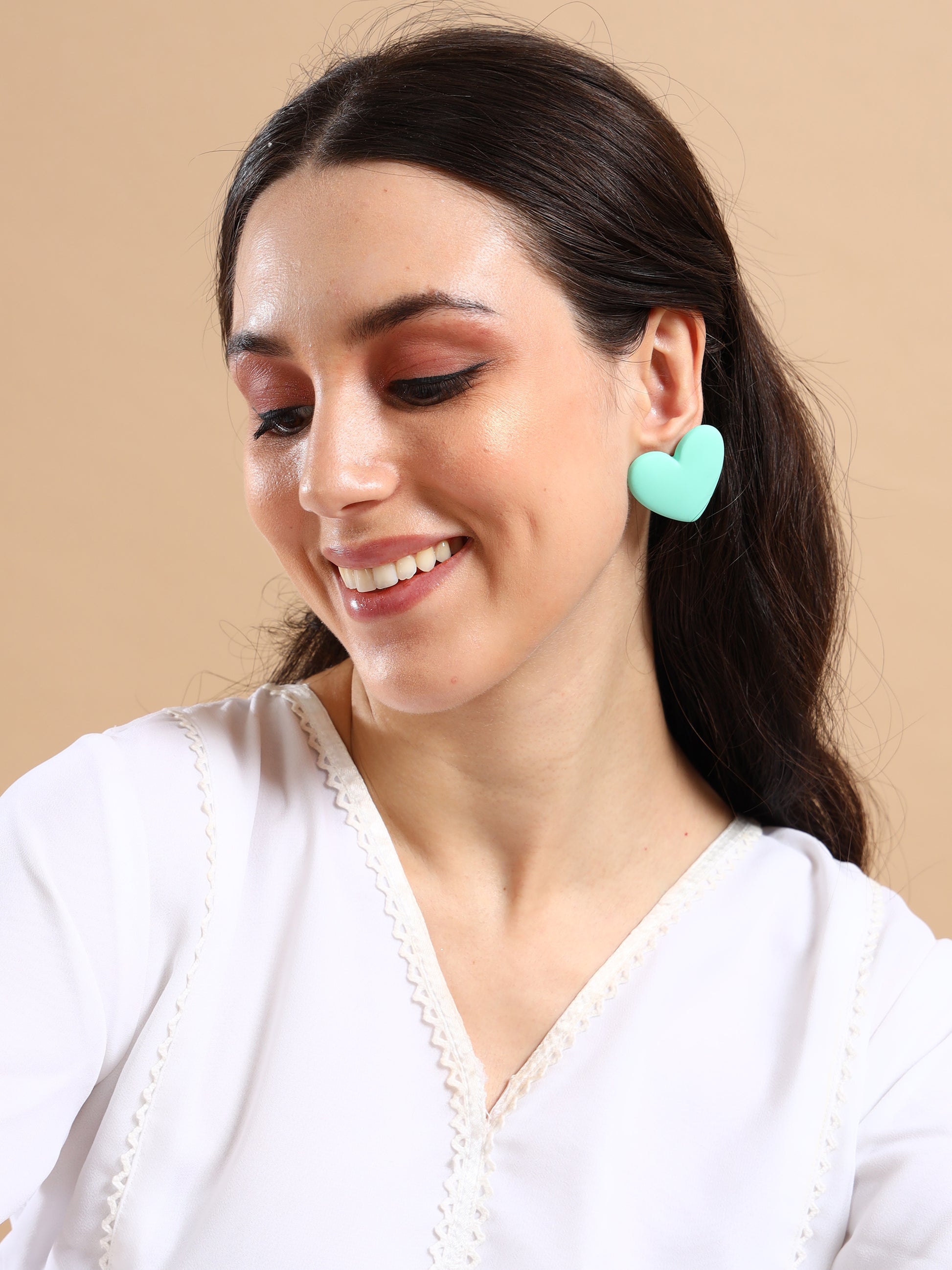 A woman with long dark hair and glowing makeup smiles in a white top, showcasing the Love Hearts Earrings—large, mint-green, heart-shaped gold-plated accessories—against a beige background.