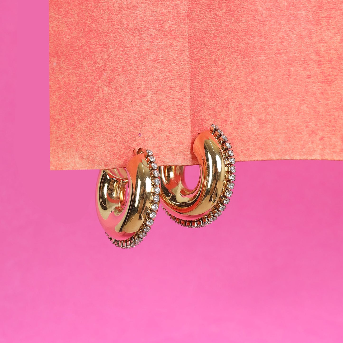 The Half Circle Hoop Earrings, finished in 24K gold plating and accented with a row of sparkling zircon stones, rest on the edge of a textured pink surface against a bright pink background.