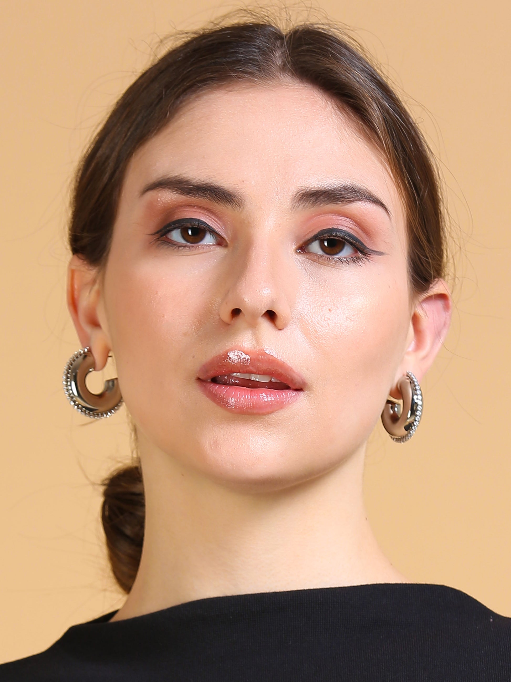 A woman with light skin and brown hair in a low bun, wearing glossy nude lipstick, subtle eye makeup, Half Circle Silver Hoop earrings, and a black top poses against a plain beige background.