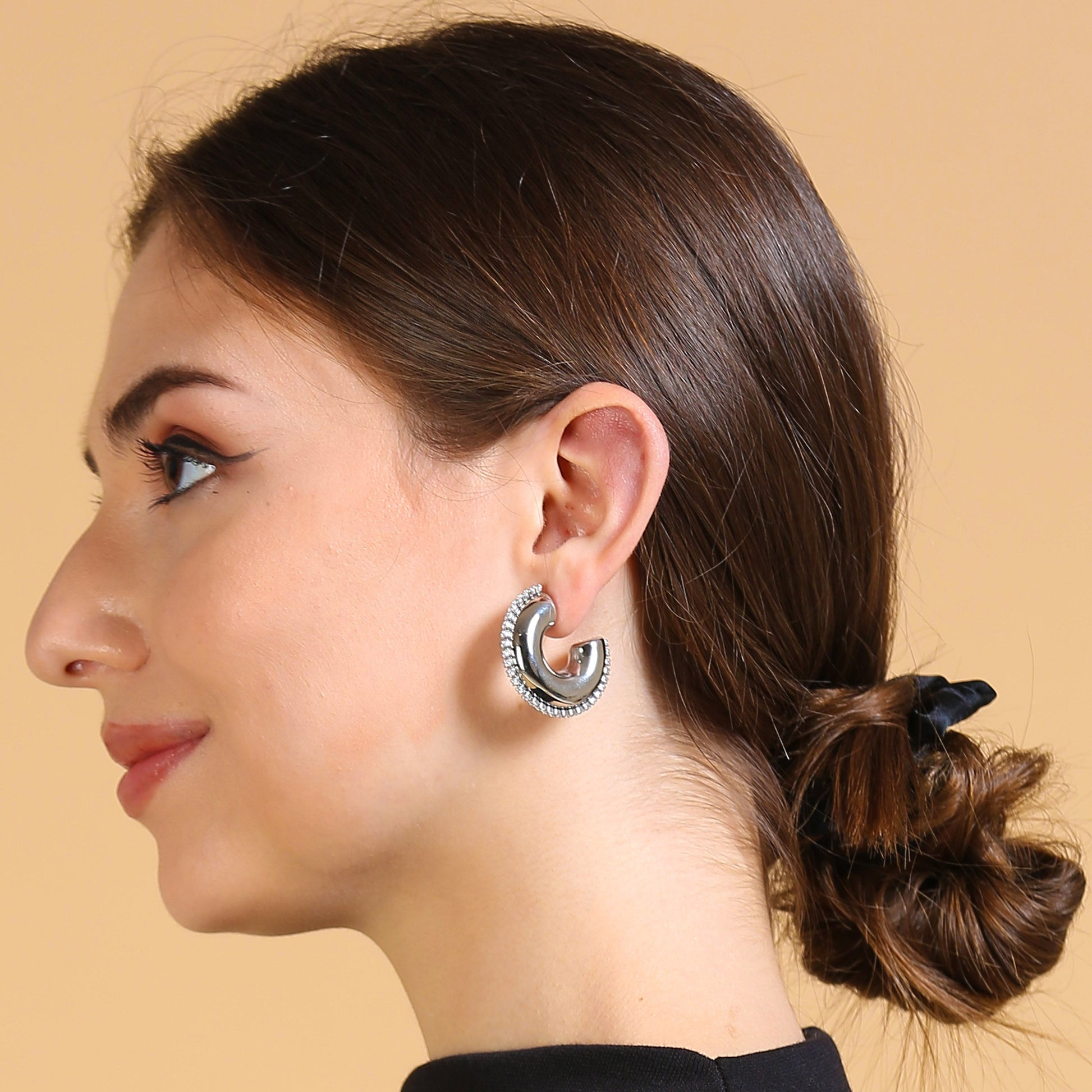 A woman with brown hair in a low bun, wearing a black top and the Half Circle Silver Hoop earrings with a shiny silver finish, smiles slightly and looks to the left against a beige background.