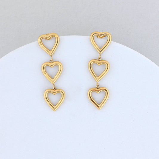 The Gold Heart Dangler Earrings feature three linked open heart shapes in each earring, beautifully displayed against a light grey and white background.