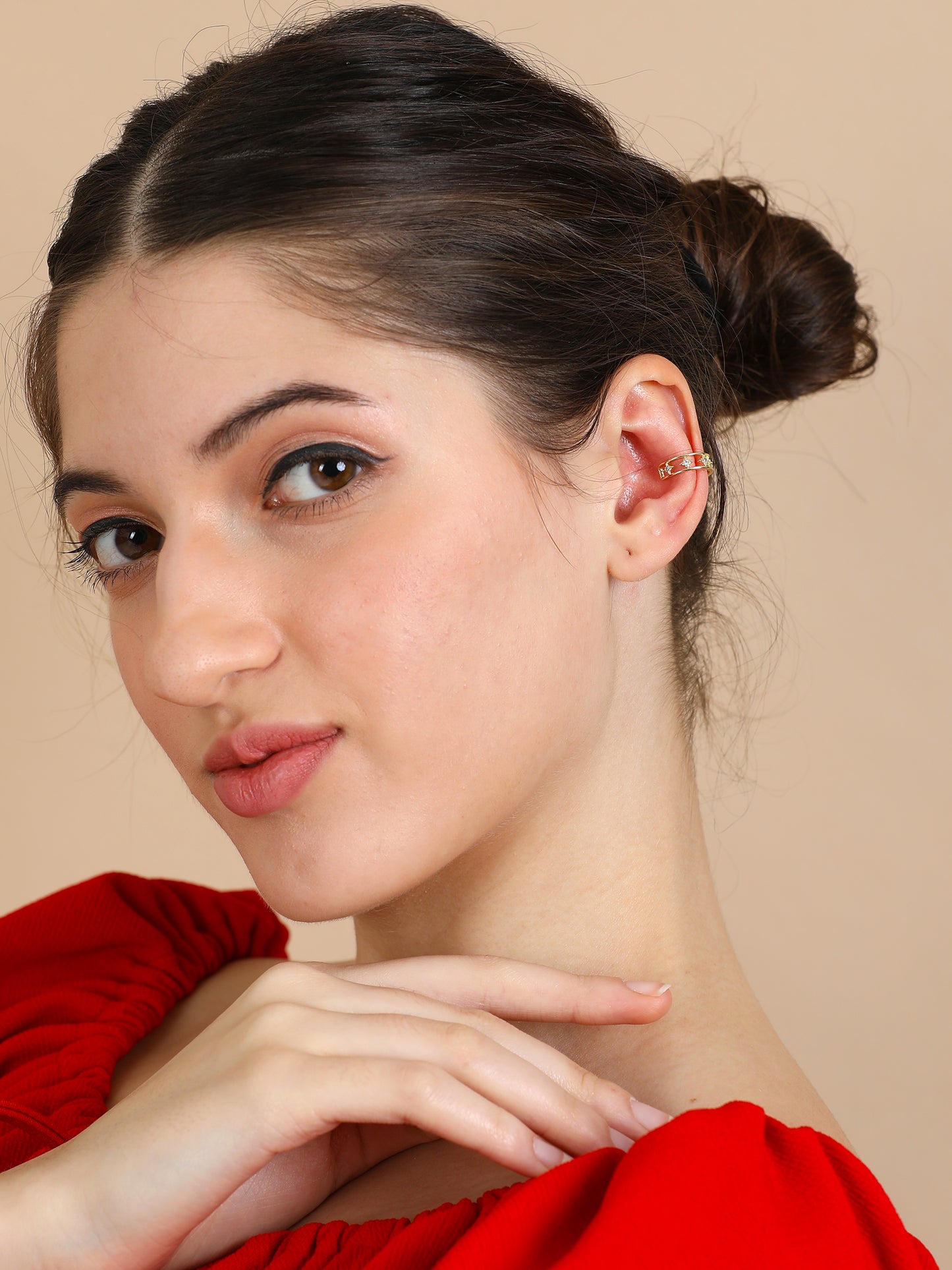 A fair-skinned woman with dark hair in a bun wears a red top and smiles softly at the camera, highlighting the Small Gold Ear Cuff on her left ear as she gently touches her chin.
