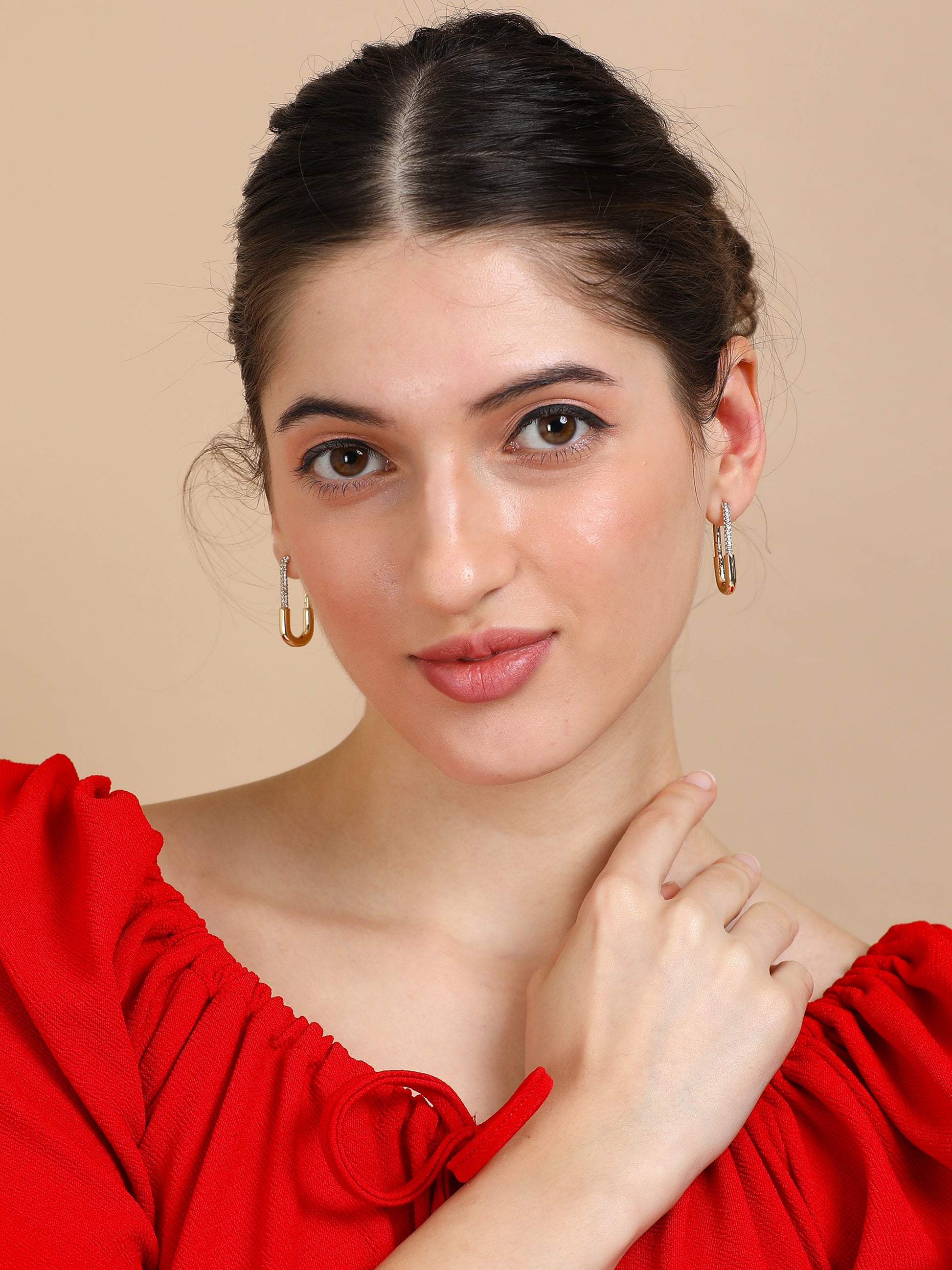 A woman with fair skin and dark hair tied back wears Long Rectangle Gold Hoops and a red off-shoulder top, gazing neutrally at the camera while gently touching her neck.