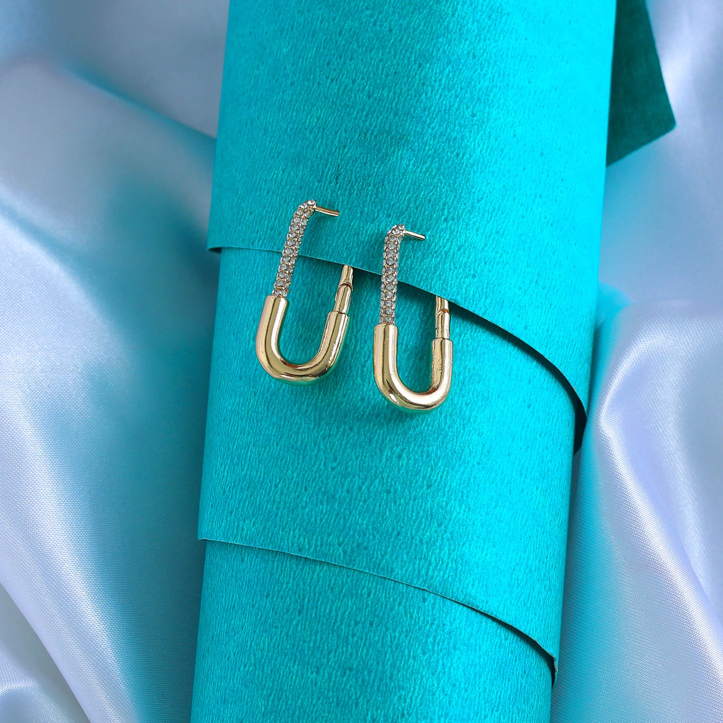 Long Rectangle Gold Hoops with textured upper sections rest on a rolled teal sheet, surrounded by shiny, light blue satin fabric.