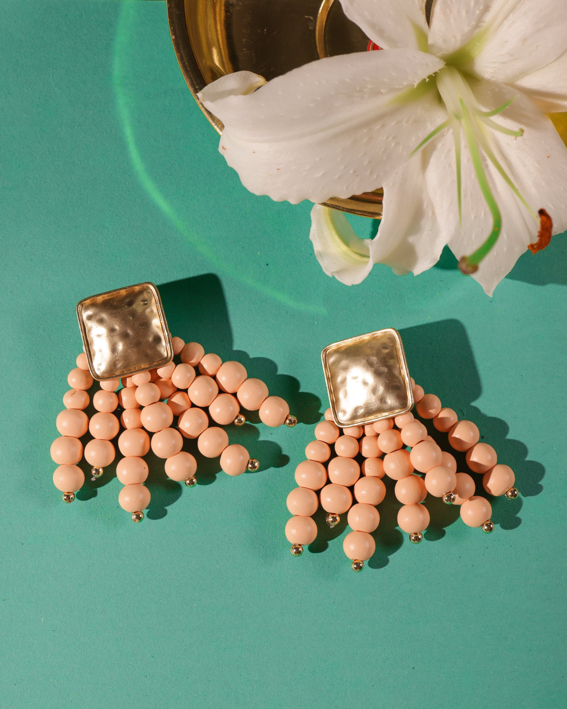 Beady Peach and Gold Earrings with gold-plated square tops and hanging peach beads are shown on a teal surface beside a white lily and gold dish—perfect for adding luxury vibes to your summer jewelry collection.