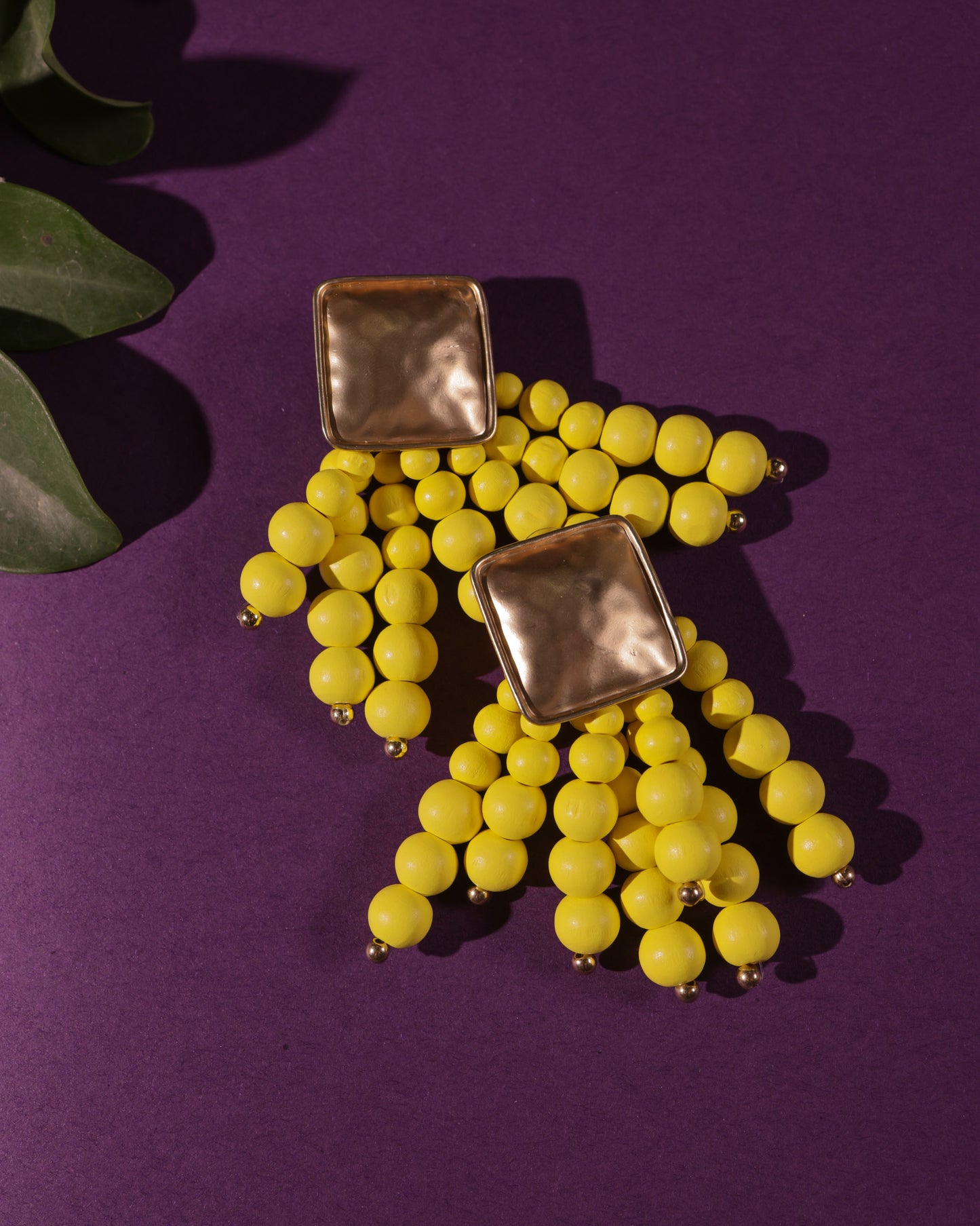 Beady Yellow and Gold Earrings featuring gold-plated square studs with dangling yellow bead clusters, perfect for summer, shown on a purple background with a green leaf in the corner.