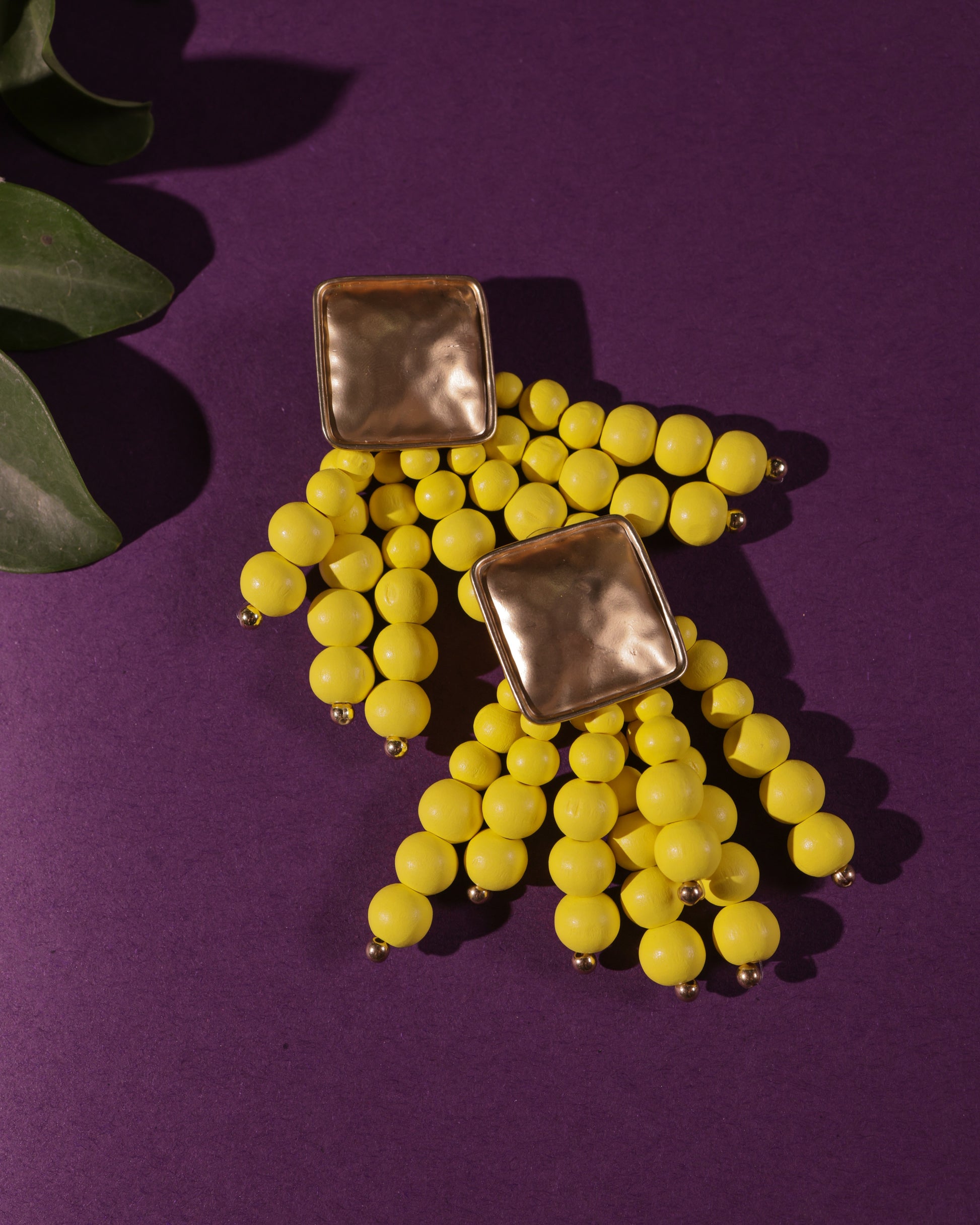 Beady Yellow and Gold Earrings featuring gold-plated square studs with dangling yellow bead clusters, perfect for summer, shown on a purple background with a green leaf in the corner.