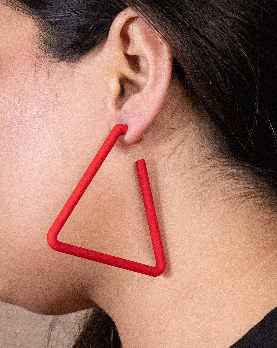 Close-up of a person's ear wearing Triangle Earrings—large, matte red, and open triangle-shaped. This affordable luxury piece pops against a neutral background, with dark hair tucked neatly behind the ear.