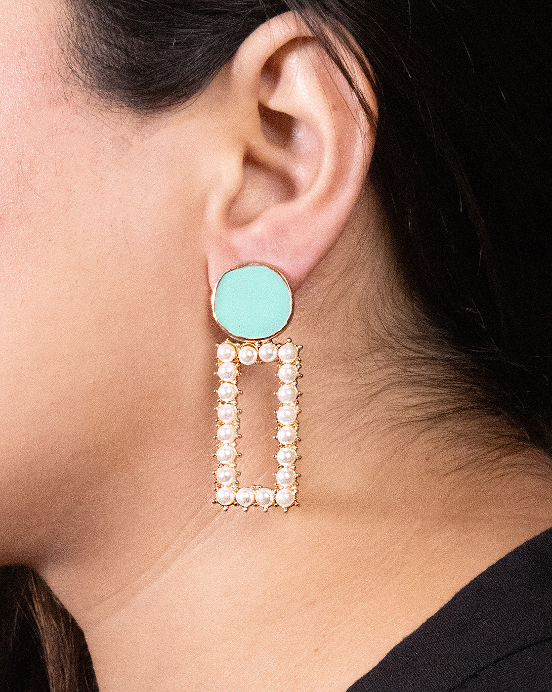 A close-up of a woman wearing the Teal a Pearl Earrings: gold-plated statement pieces with a round mint-green stud on brass alloy and a large rectangular drop adorned with small pearls.