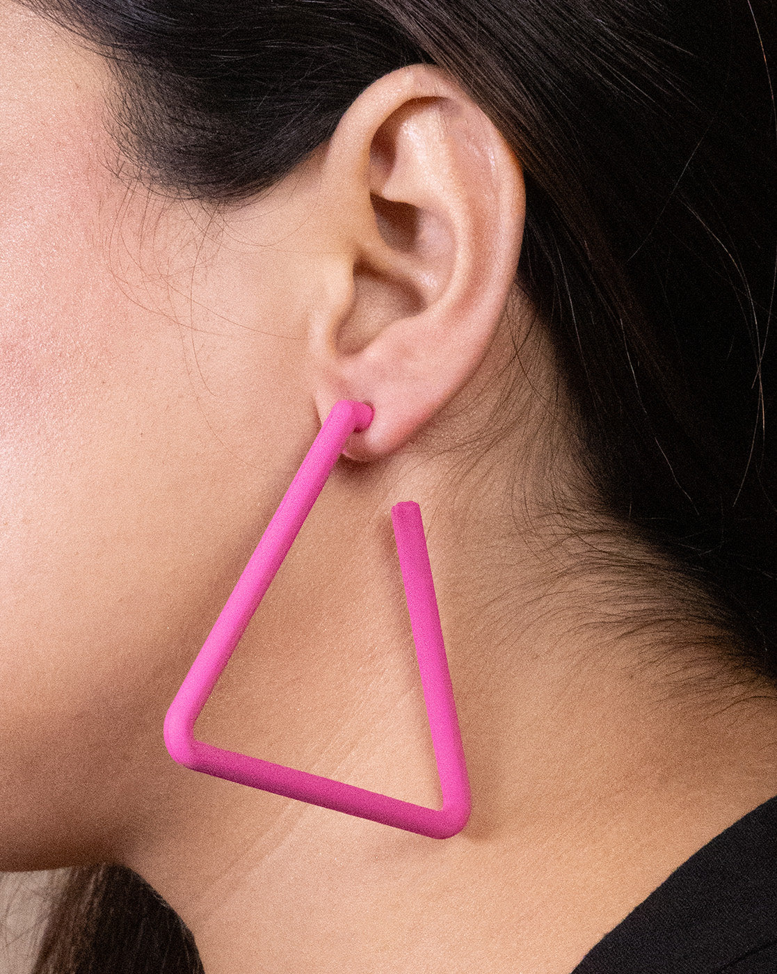 Close-up of a person's ear wearing Triangle Earrings—large, bold, geometric triangles in bright pink with 18K gold plating. Hair pulled back and black top highlight the affordable luxury of these statement earrings.