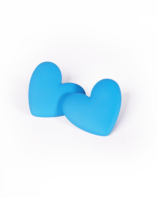 Two Love Hearts Earrings, blue and resembling lab-created stones, are closely positioned on a white background with one heart slightly overlapping the other.