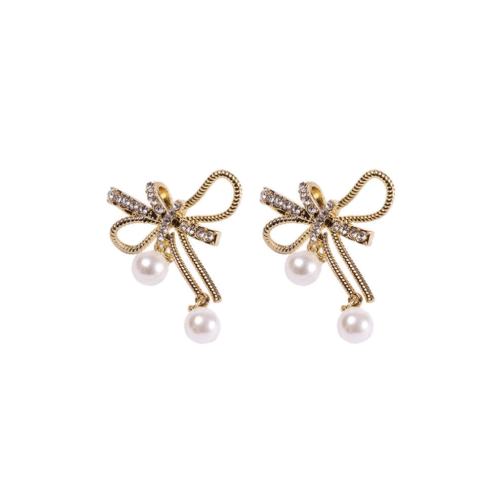 The Bow & Pearl Earrings feature 24K gold plating, bow shapes adorned with rhinestones, and a dangling white pearl on each earring, all inspired by elegant Korean design and shown against a white background.