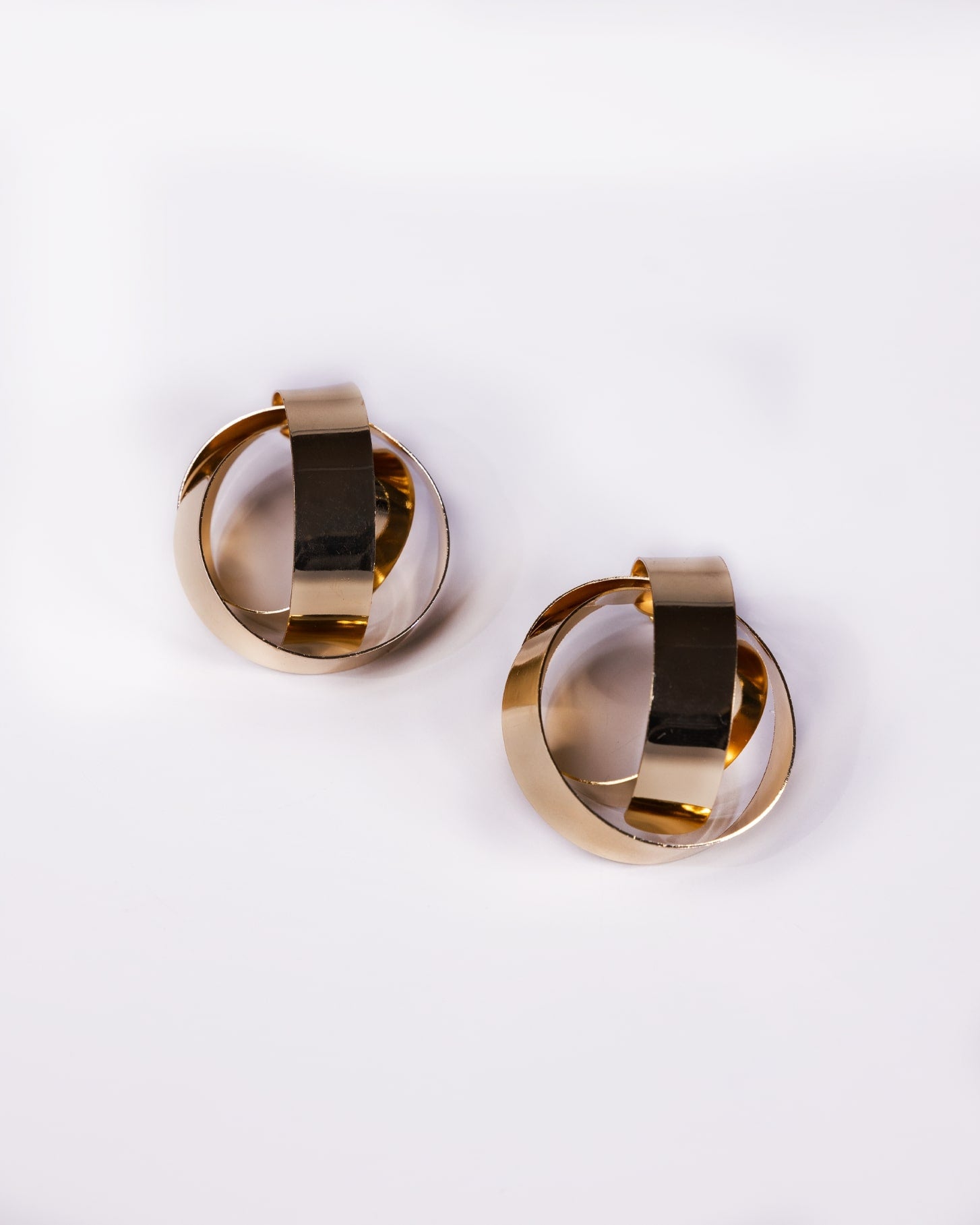 The Gold Criss Cross Earrings are 24K gold-plated, featuring interlocking circular hoops, shown on a white background.