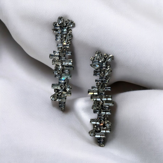 Metallic crystal earrings featuring a silver-tone, crystal-encrusted cluster design with subtle metallic edges are showcased on smooth white fabric. Their modern geometric arrangement of sparkling stones adds striking brilliance.