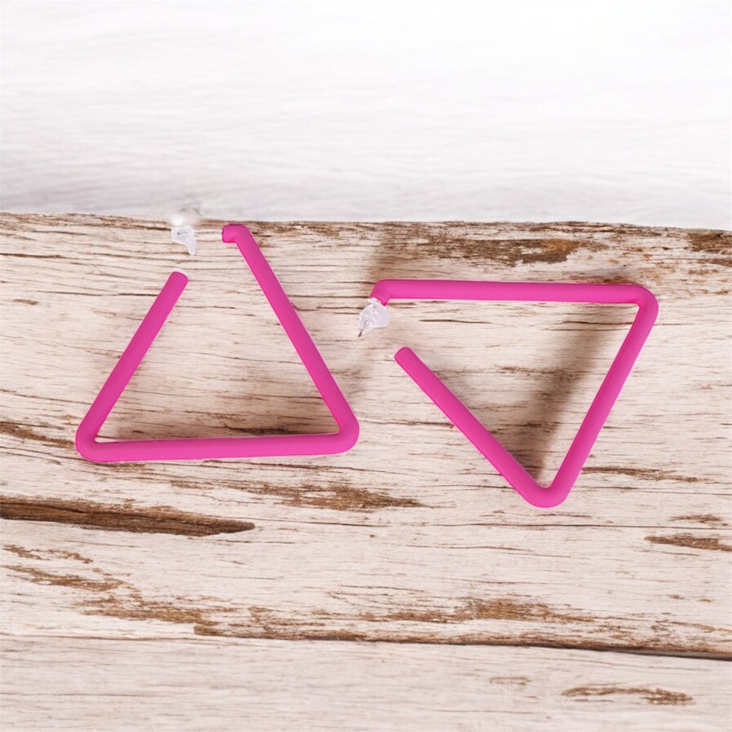 The Triangle Earrings feature a simple, open-ended geometric design in pink, forming incomplete triangles. Resting on a wooden surface with a white background, these affordable luxury earrings add modern elegance to any look.