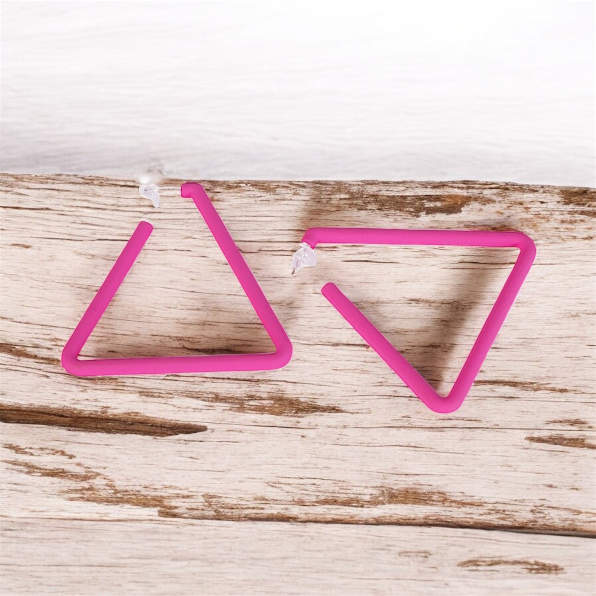 The Triangle Earrings feature a simple, open-ended geometric design in pink, forming incomplete triangles. Resting on a wooden surface with a white background, these affordable luxury earrings add modern elegance to any look.