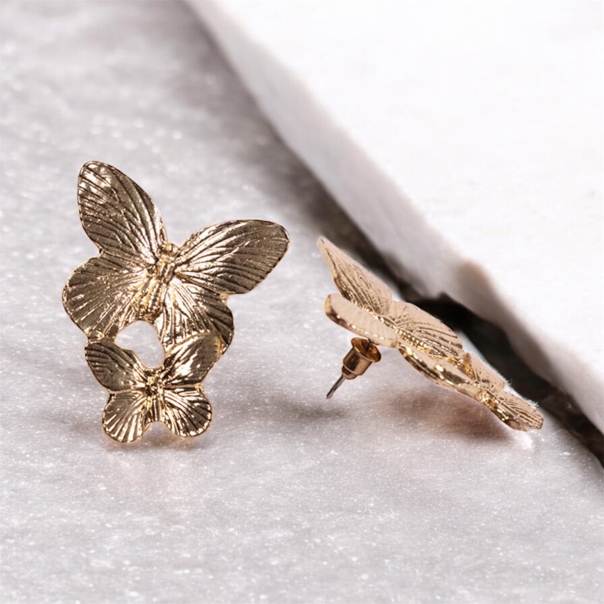 The Butterfly Earrings, a pair of 24K gold plated butterfly-shaped studs, are shown on a white marble surface—one lying flat and the other upright to highlight their intricate details and texture.