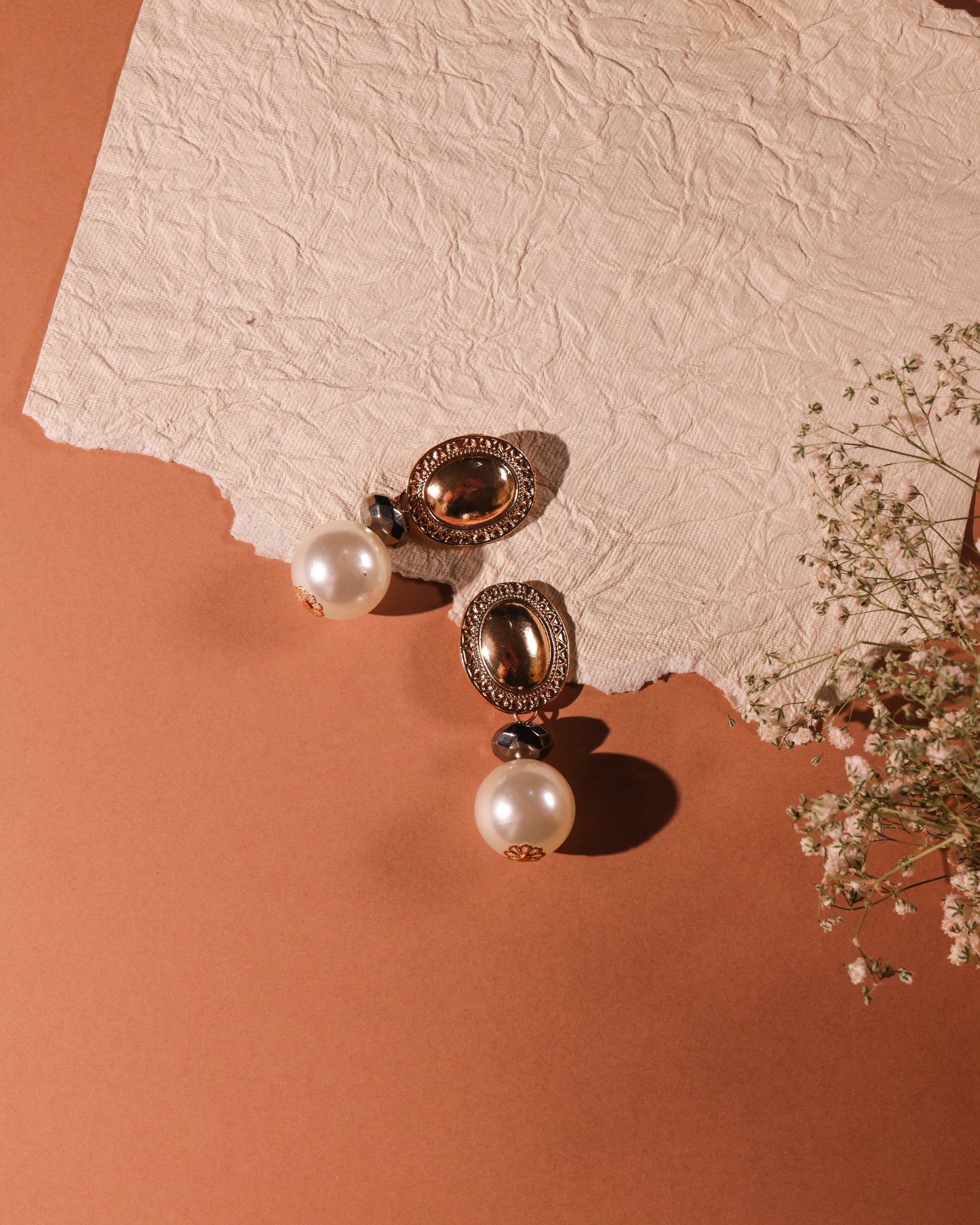 The Pearl & Shield Earrings feature brass alloy oval settings with black stone accents and large white pearls, displayed on textured paper and a peach surface beside delicate white baby's breath flowers.