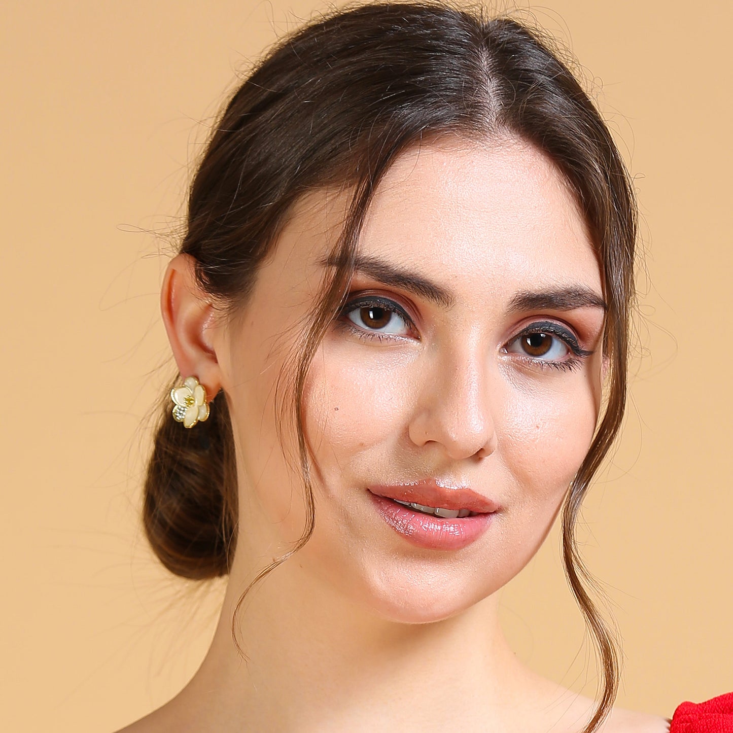 A woman with fair skin and brown hair in a low bun, wearing enamel flower earrings and natural makeup, stands against a solid beige background. She smiles softly, with loose strands framing her face.