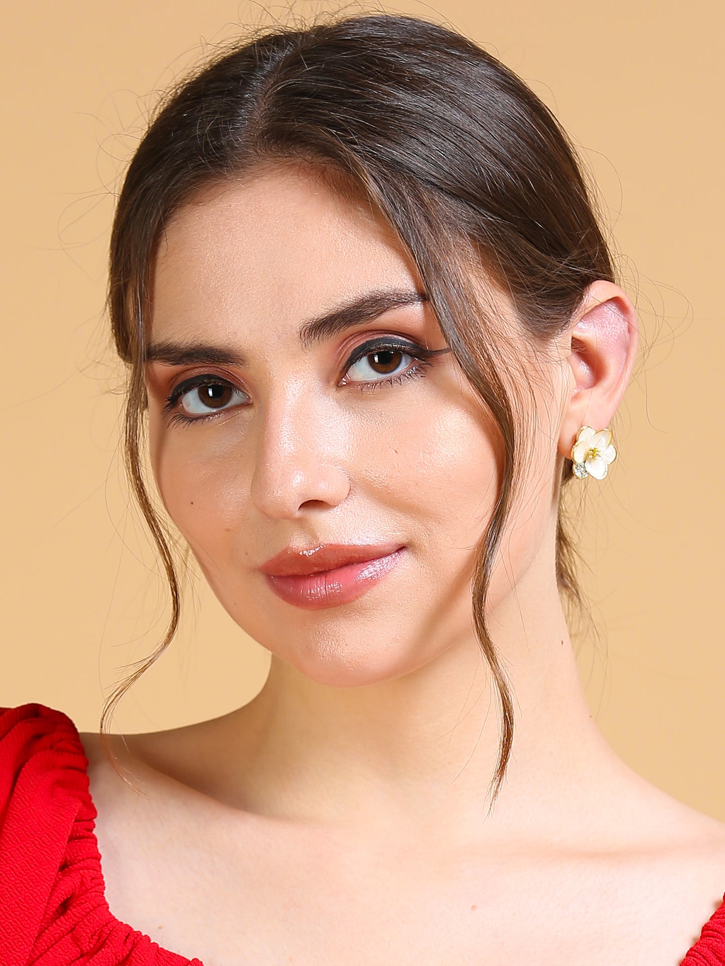A woman with long brown hair in a low bun wears Enamel flower earrings and a red top, posing against a beige background. She has natural makeup with soft tendrils framing her face.